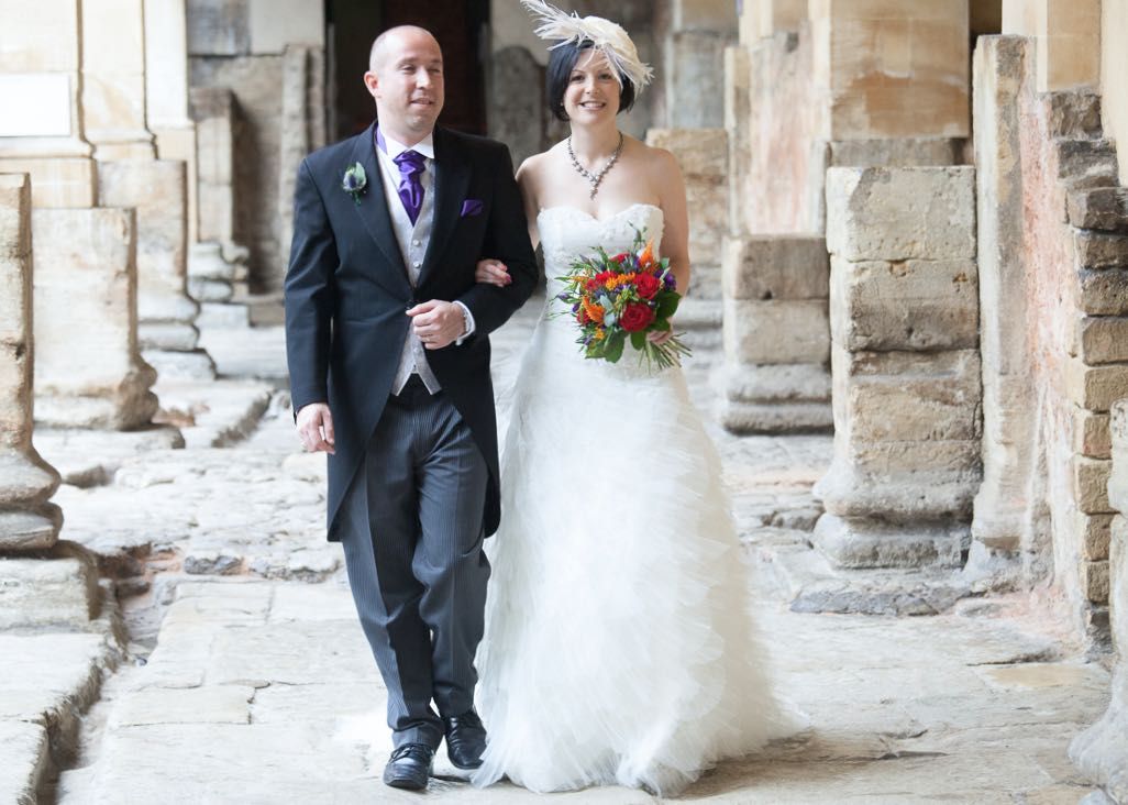 A couple walks arm in arm through a historic stone colonnade on their wedding day, with the bride holding red flowers.