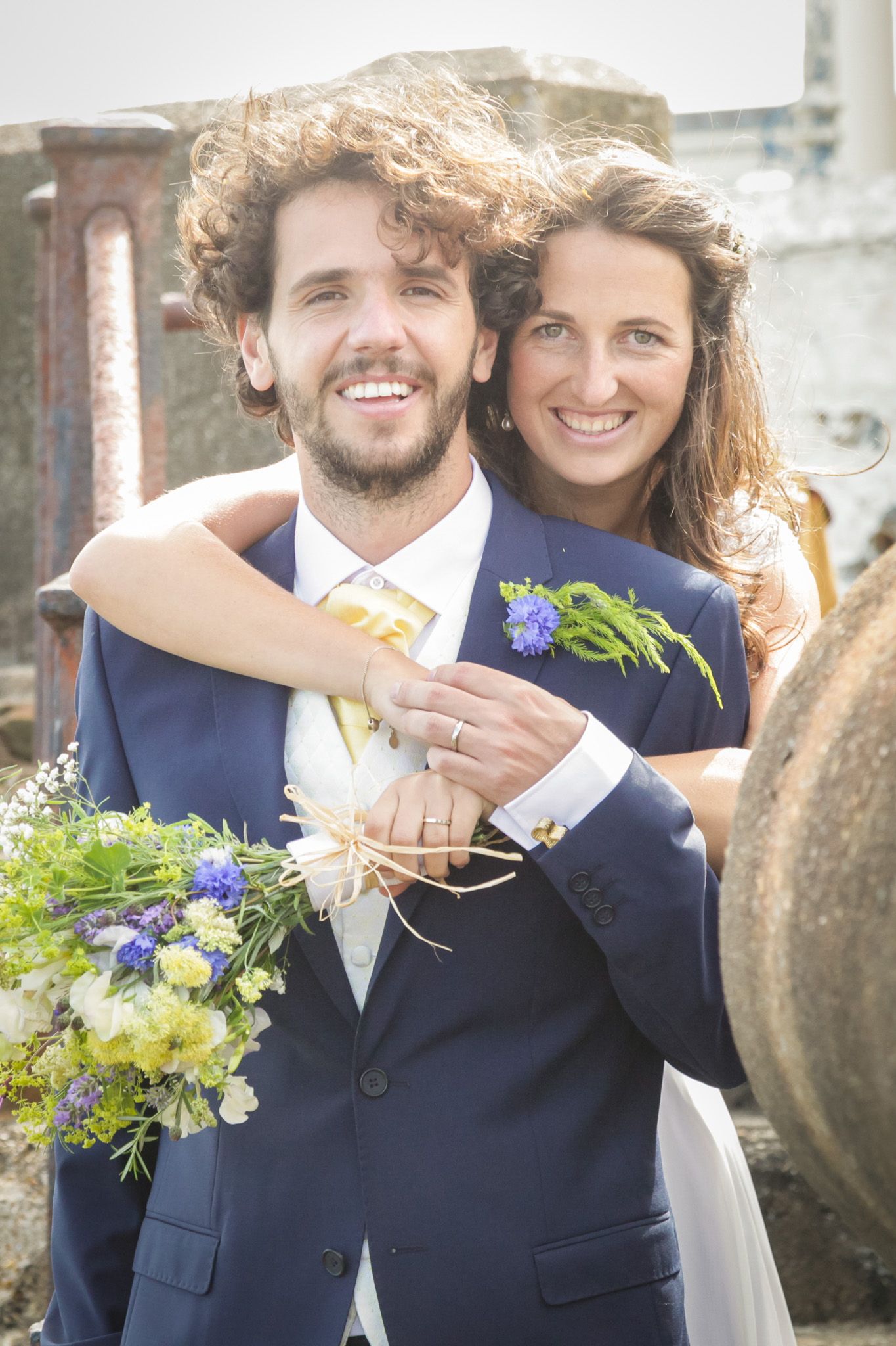 Joyful wedding couple embraces outdoors with blue and white floral bouquet details.