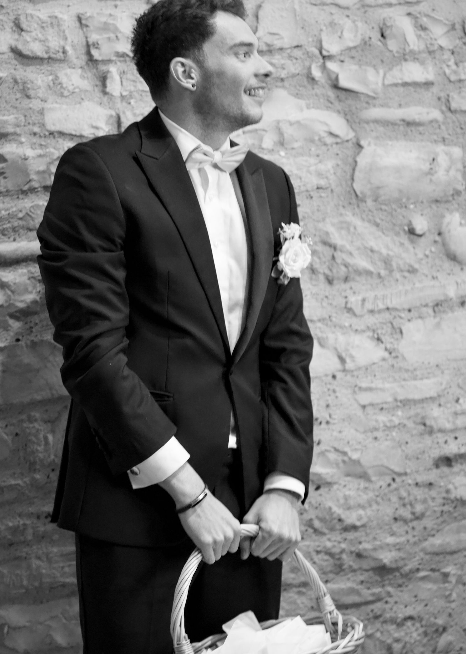 Black and white portrait of a groom holding a basket against a stone wall.