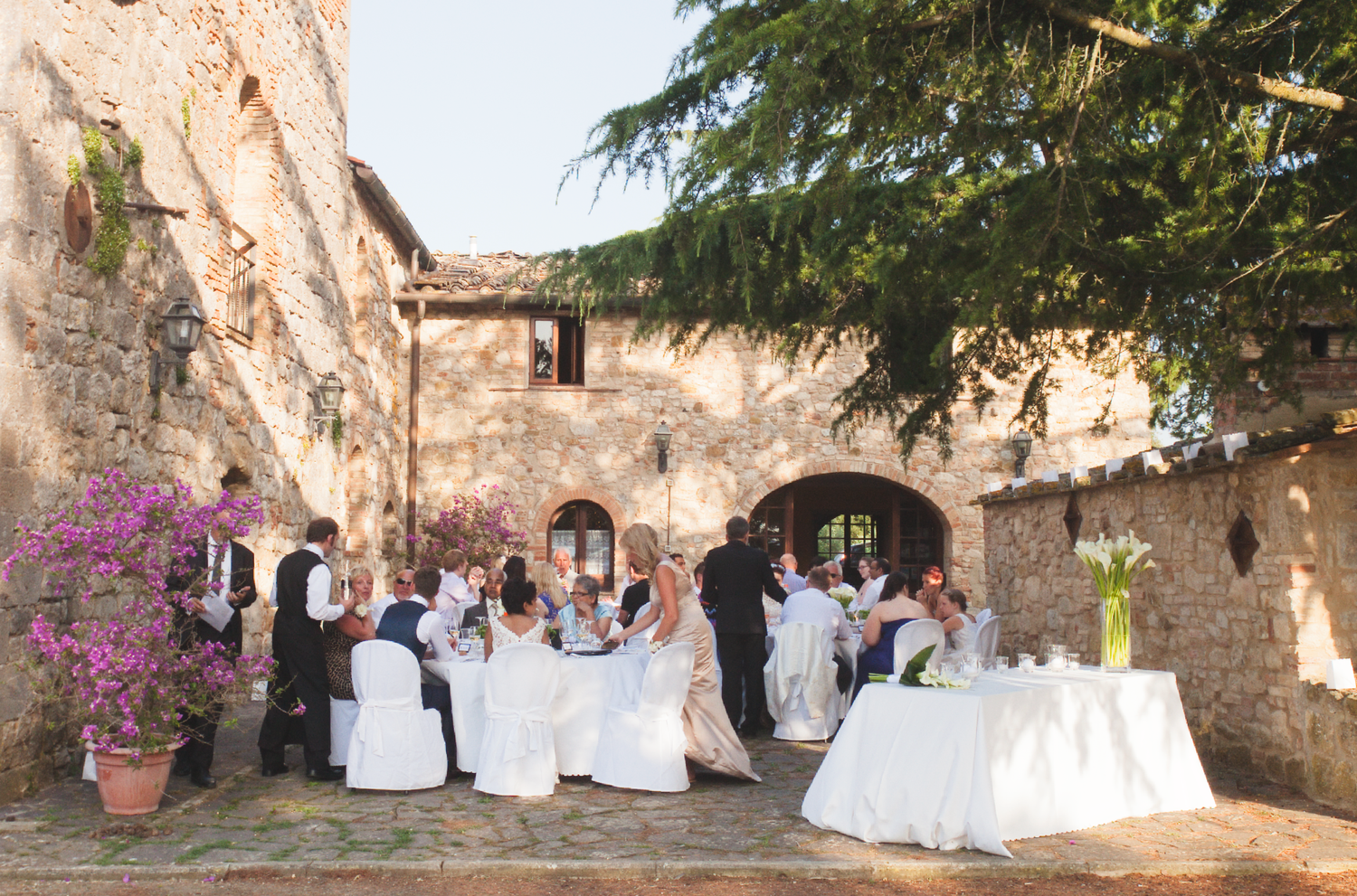 Wedding guests seated at elegant outdoor tables with white linens and stone wall backdrop.