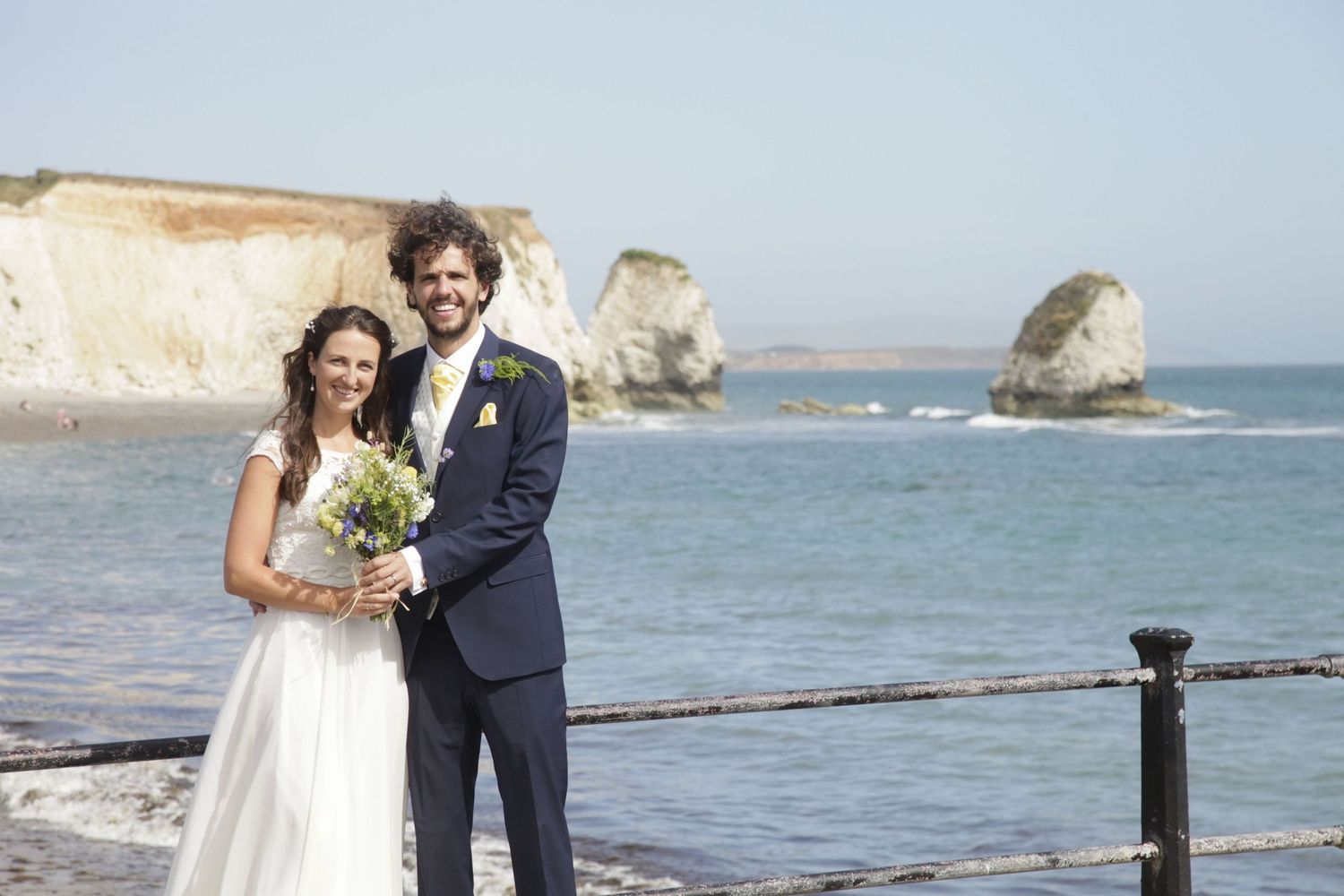 Newlyweds standing together on beach walkway with dramatic coastal scenery.