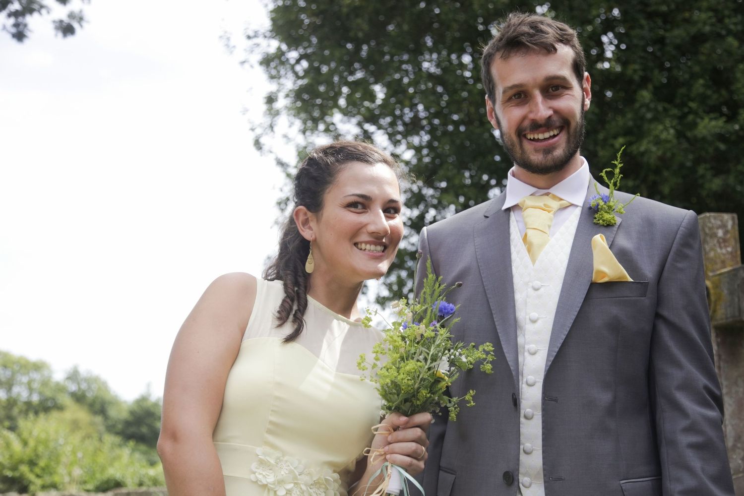 Wedding party members in formal attire pose together outdoors near trees and greenery.
