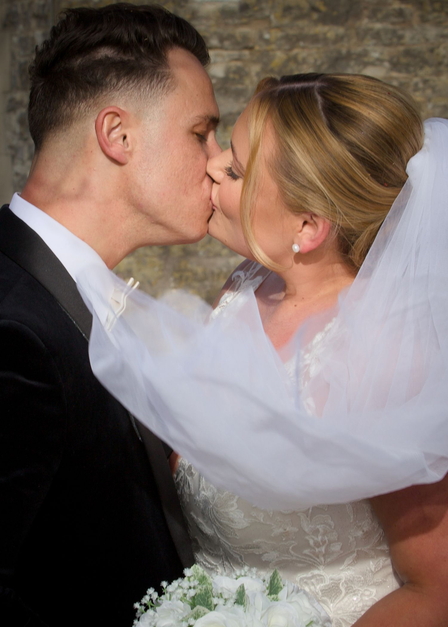 Romantic wedding kiss portraits against rustic stone wall backdrop.