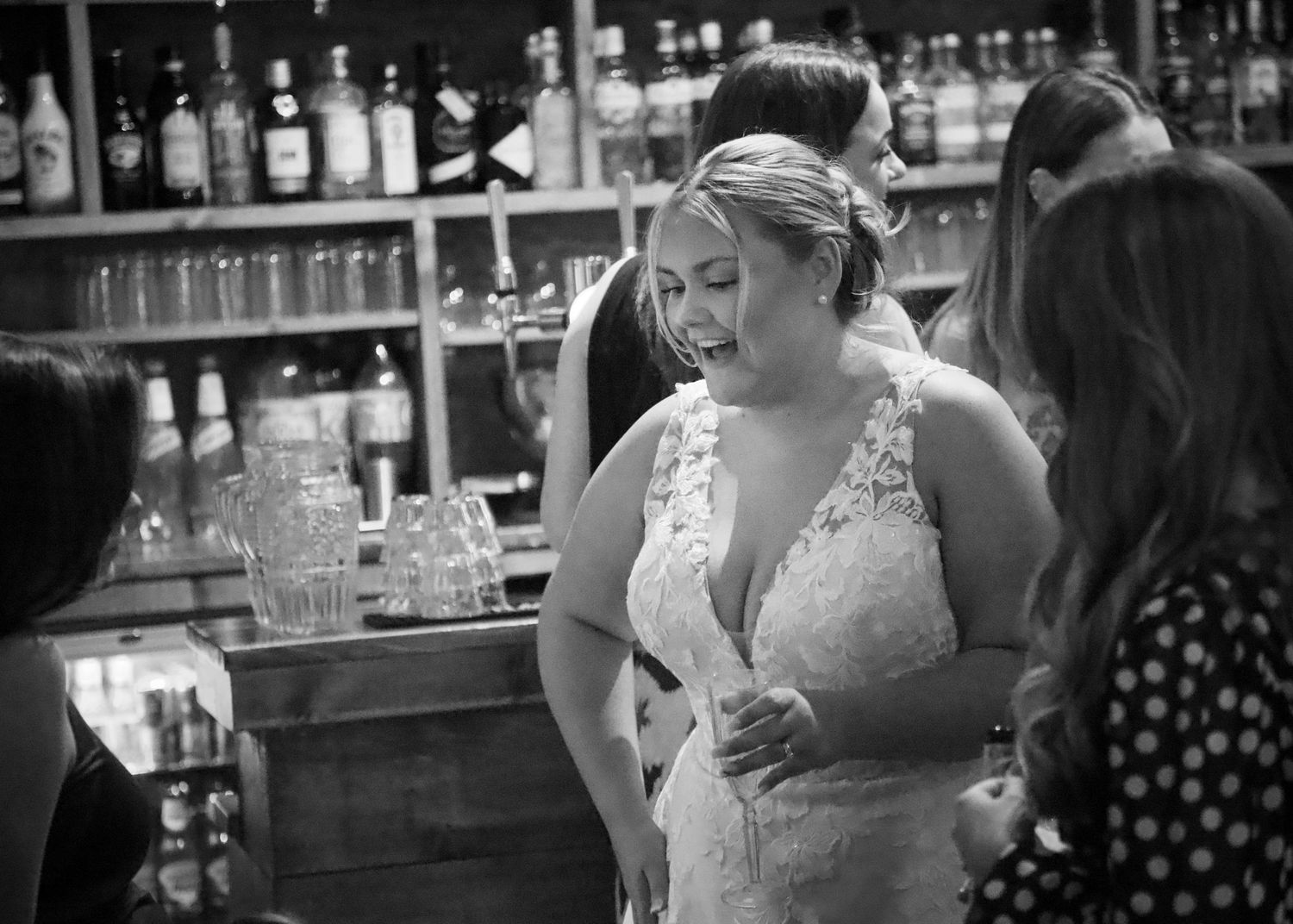 A bride in a lace dress stands at a bar with bottles displayed in the background.