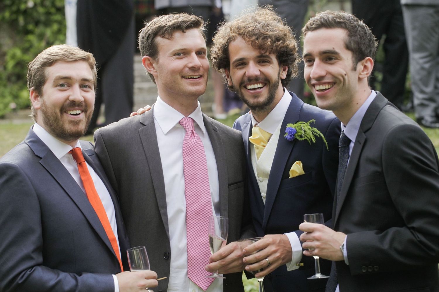 Groomsmen in formal attire sharing laughs and drinks at an outdoor wedding reception.
