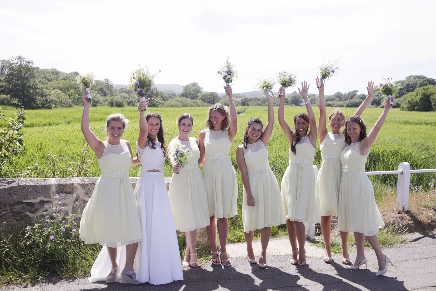 Bridesmaids in light colored dresses pose joyfully with raised arms against a scenic green landscape background.