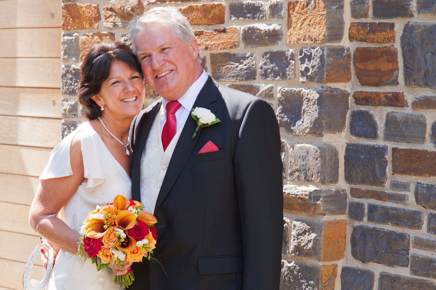 Wedding couple poses against stone wall with vibrant orange and yellow bouquet.