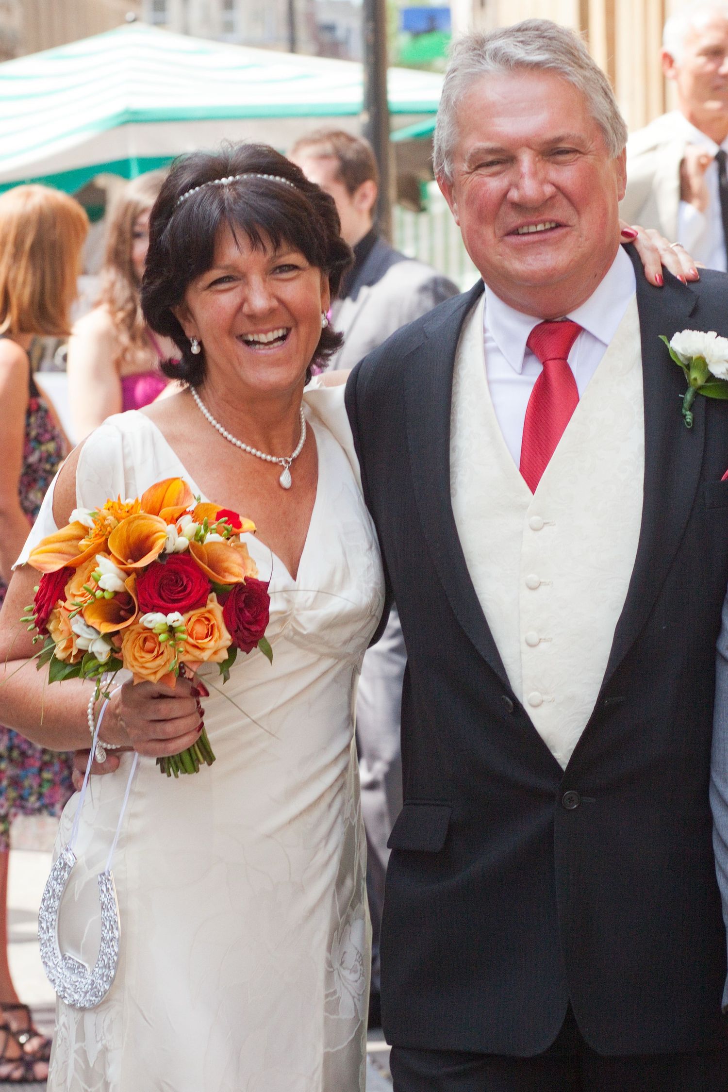 A joyful wedding couple smiling outdoors while holding a vibrant orange and red bouquet.