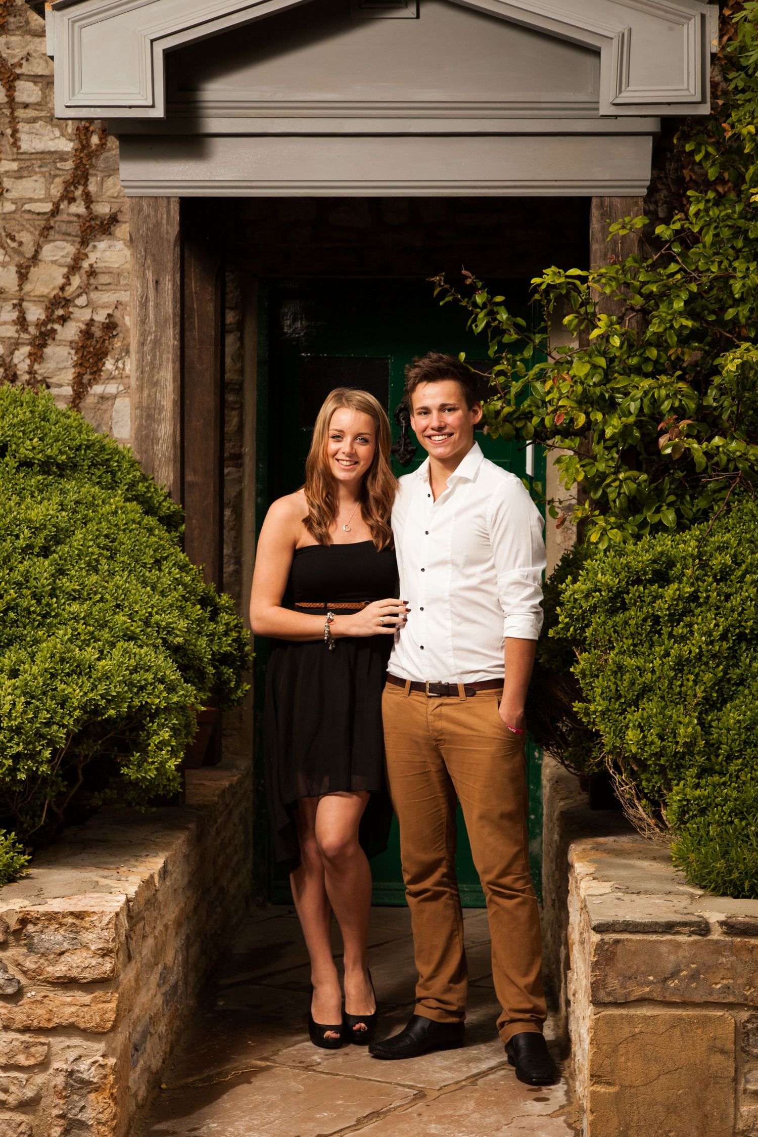 Couple poses together in front of a stone building entrance with green bushes.