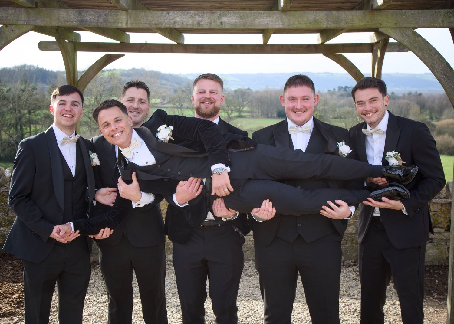Groomsmen in black suits posing playfully under a wooden pergola outdoors.