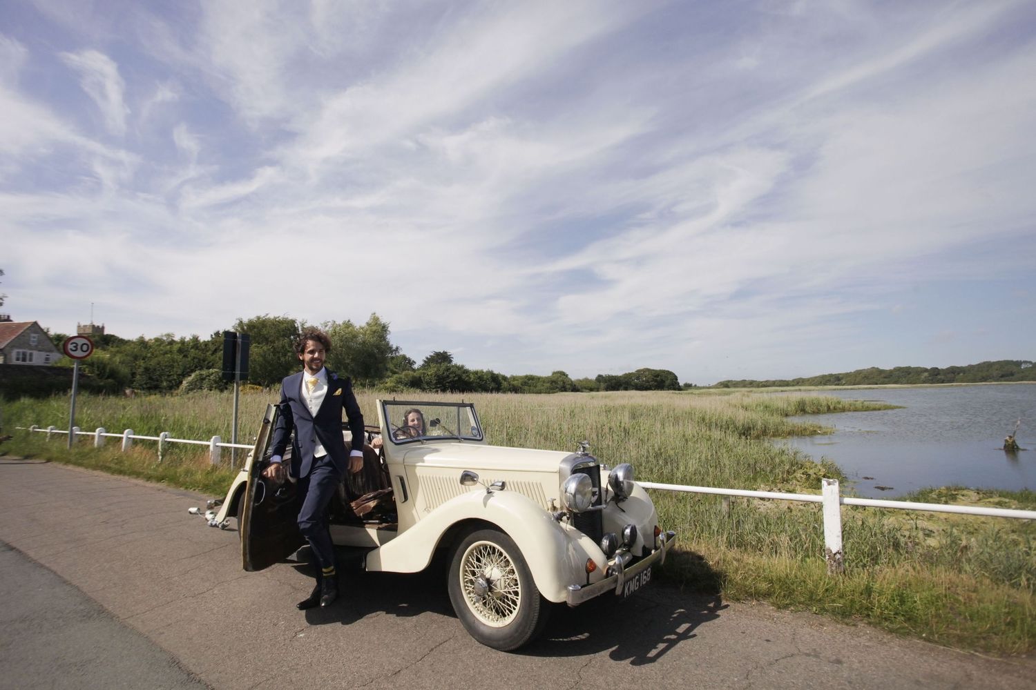 Vintage wedding car driving along coastal road with white fence and water view.