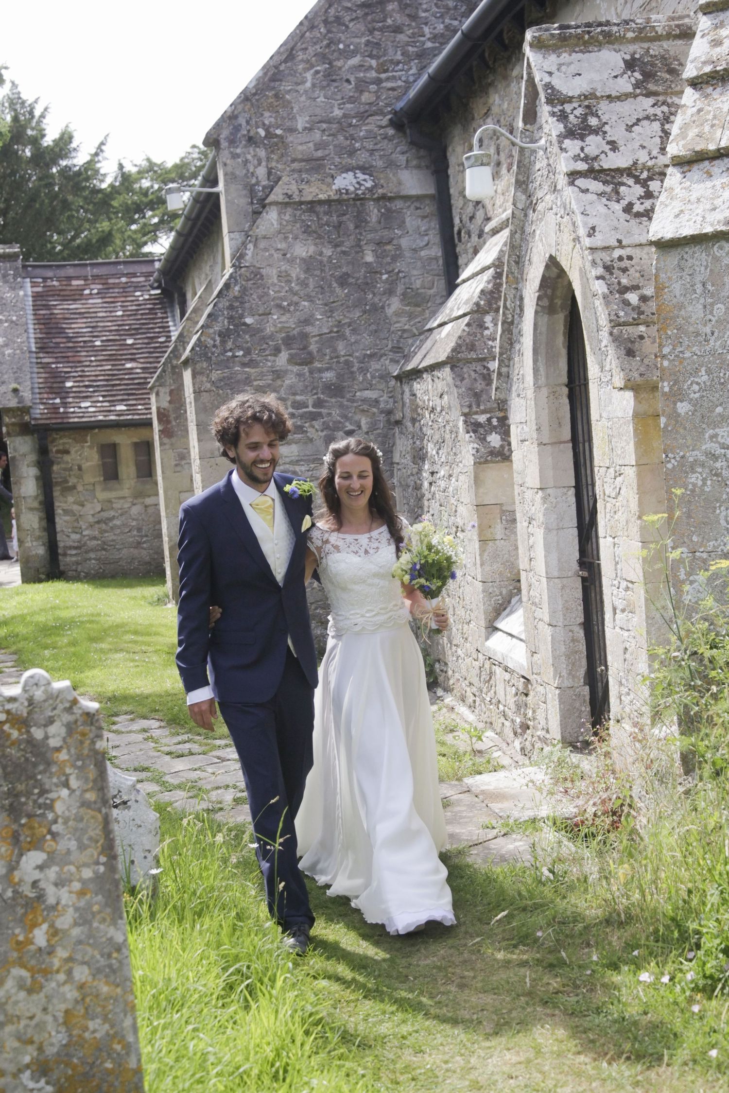 A bride and groom walk along an old stone church wall surrounded by grass and wildflowers.