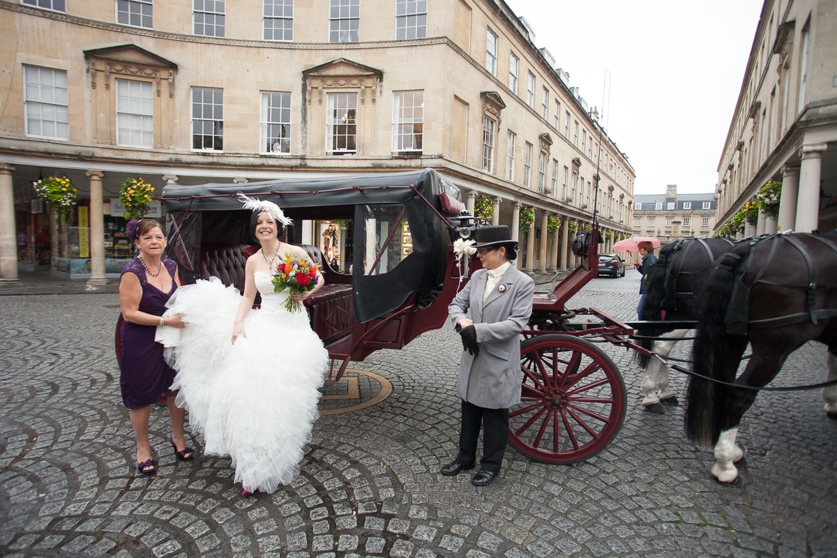 A horse-drawn carriage waits on a cobblestone street in front of historic Georgian buildings in Bath, England.