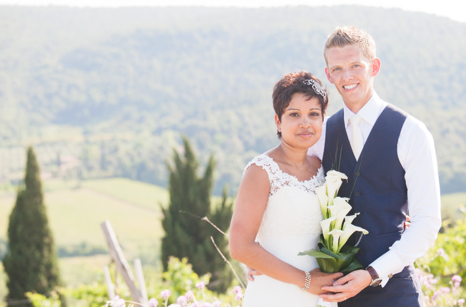 A wedding couple poses in a scenic vineyard setting with mountains and cypress trees in the background.