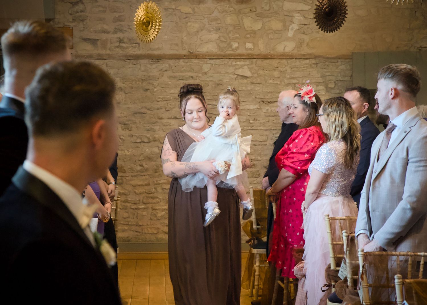 Wedding ceremony guests seated in rows of wooden chairs inside a rustic brick-walled venue.