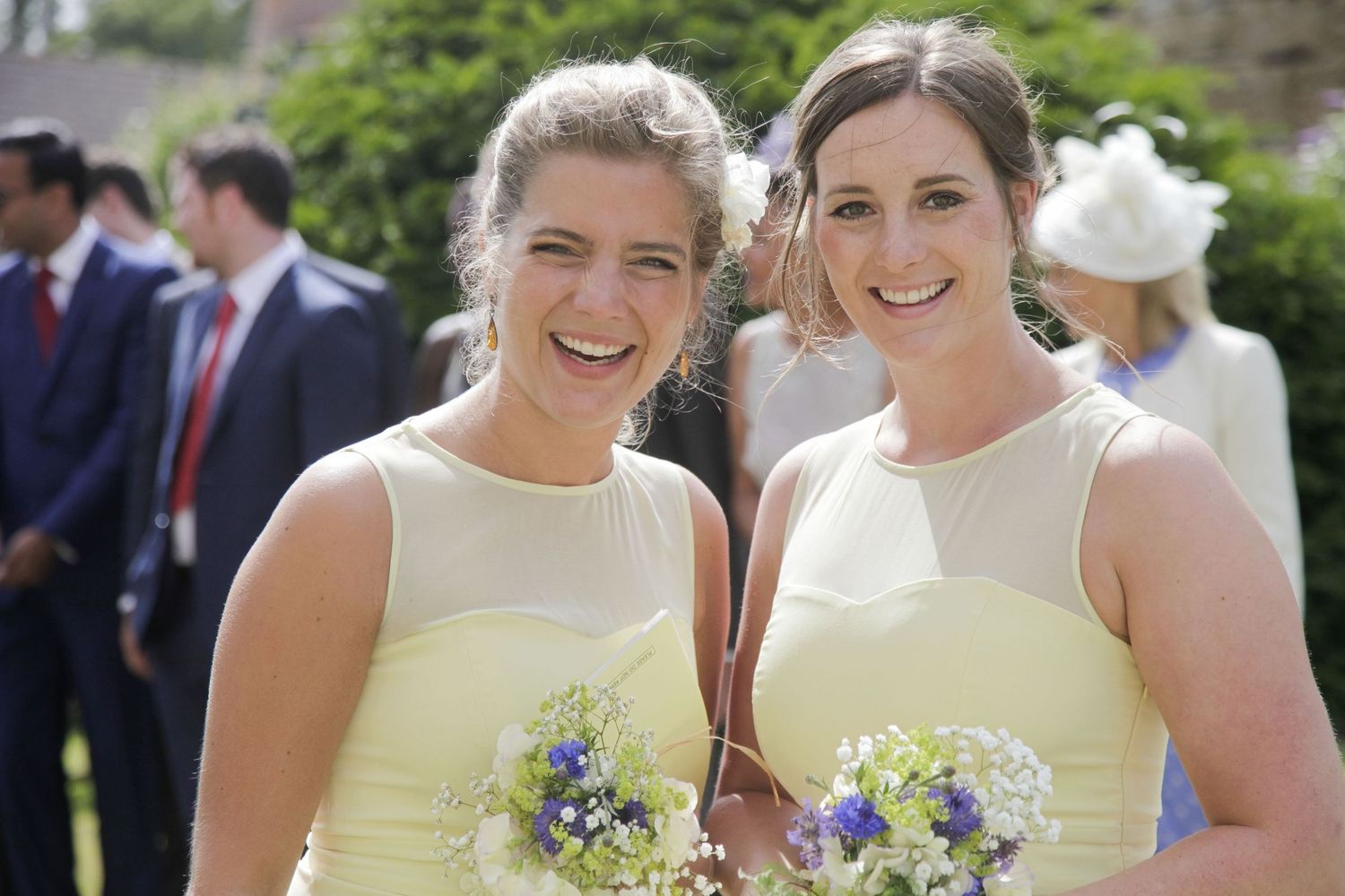 Bridesmaids in pale yellow dresses hold blue and white flower bouquets outdoors.