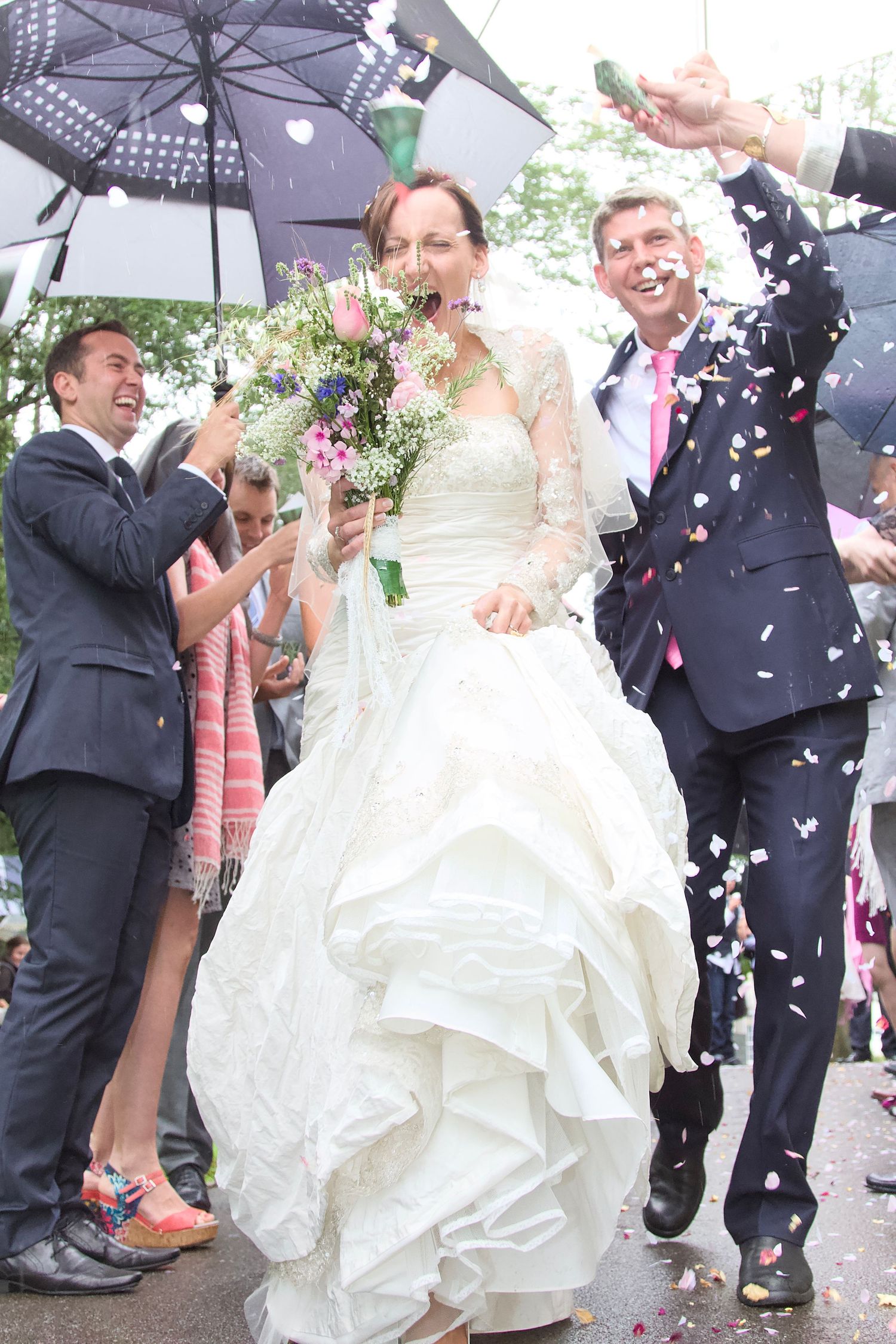 Wedding guests celebrate with umbrellas as confetti falls around the newlyweds in an outdoor celebration.