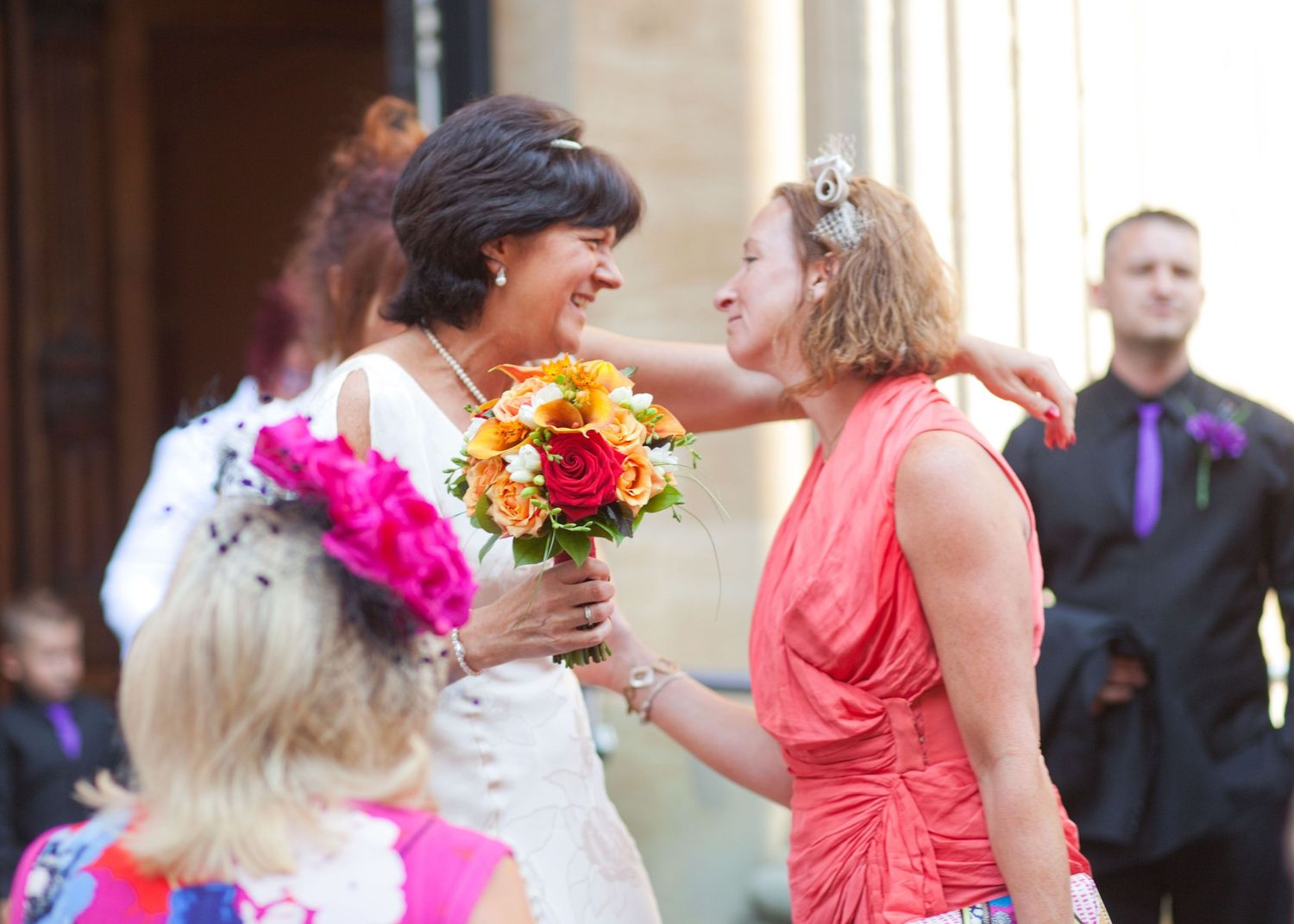 Wedding party members in coral dresses and dark suits pose together outdoors.