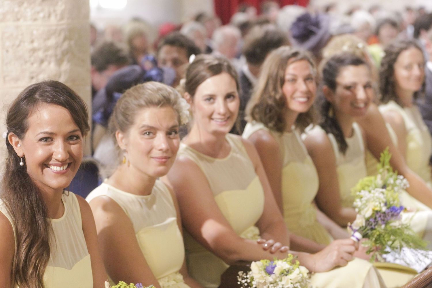 Row of bridesmaids in pale yellow dresses holding bouquets at church wedding.