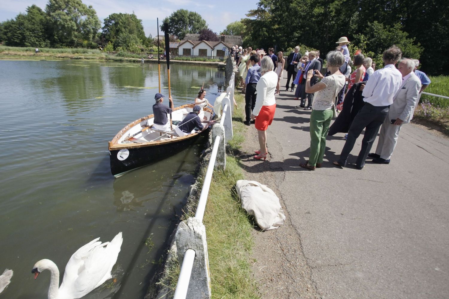 Spectators line a waterfront path near white swans as a wooden boat approaches the shore.