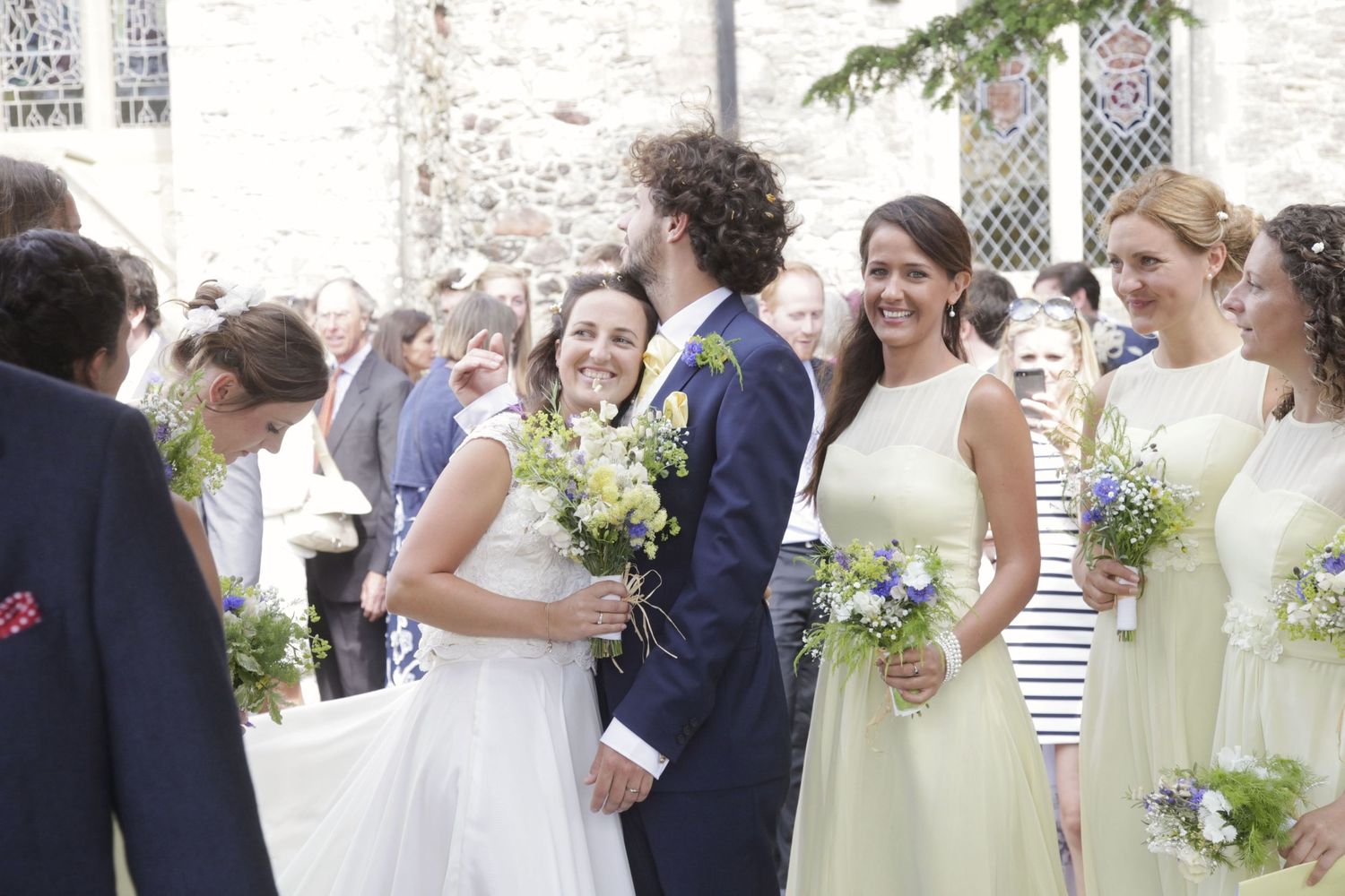 Wedding party members in pastel dresses gathered outside stone church building.