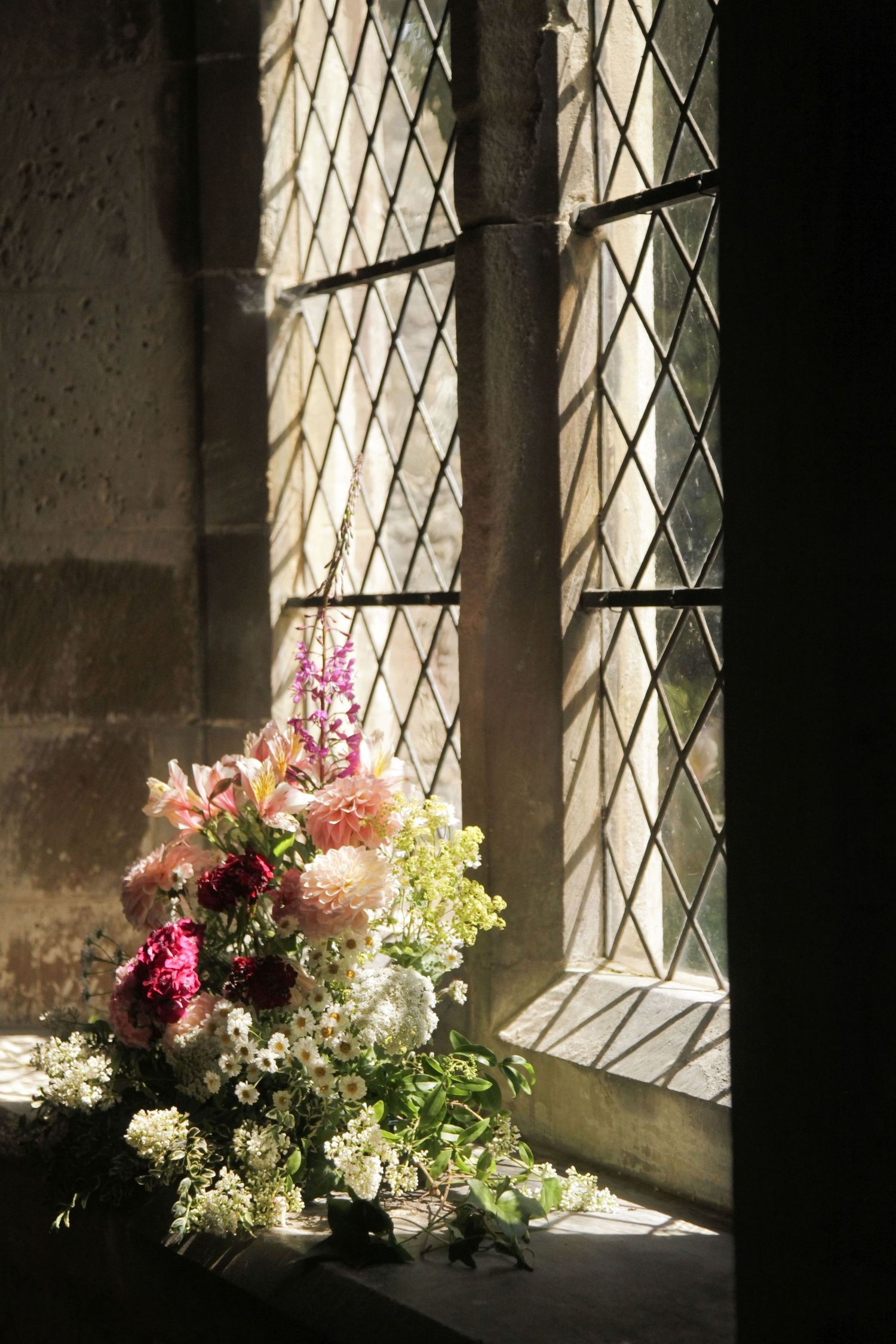 Sunlit floral arrangement by church window with pink lilies and delicate white flowers.