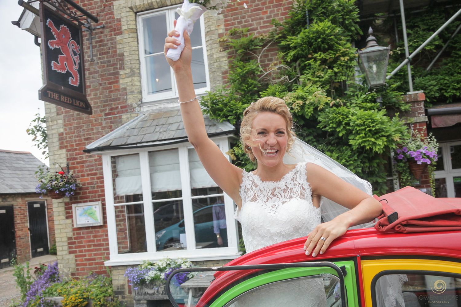 A bride waves joyfully while posing with a colorful vintage car outside a brick building.