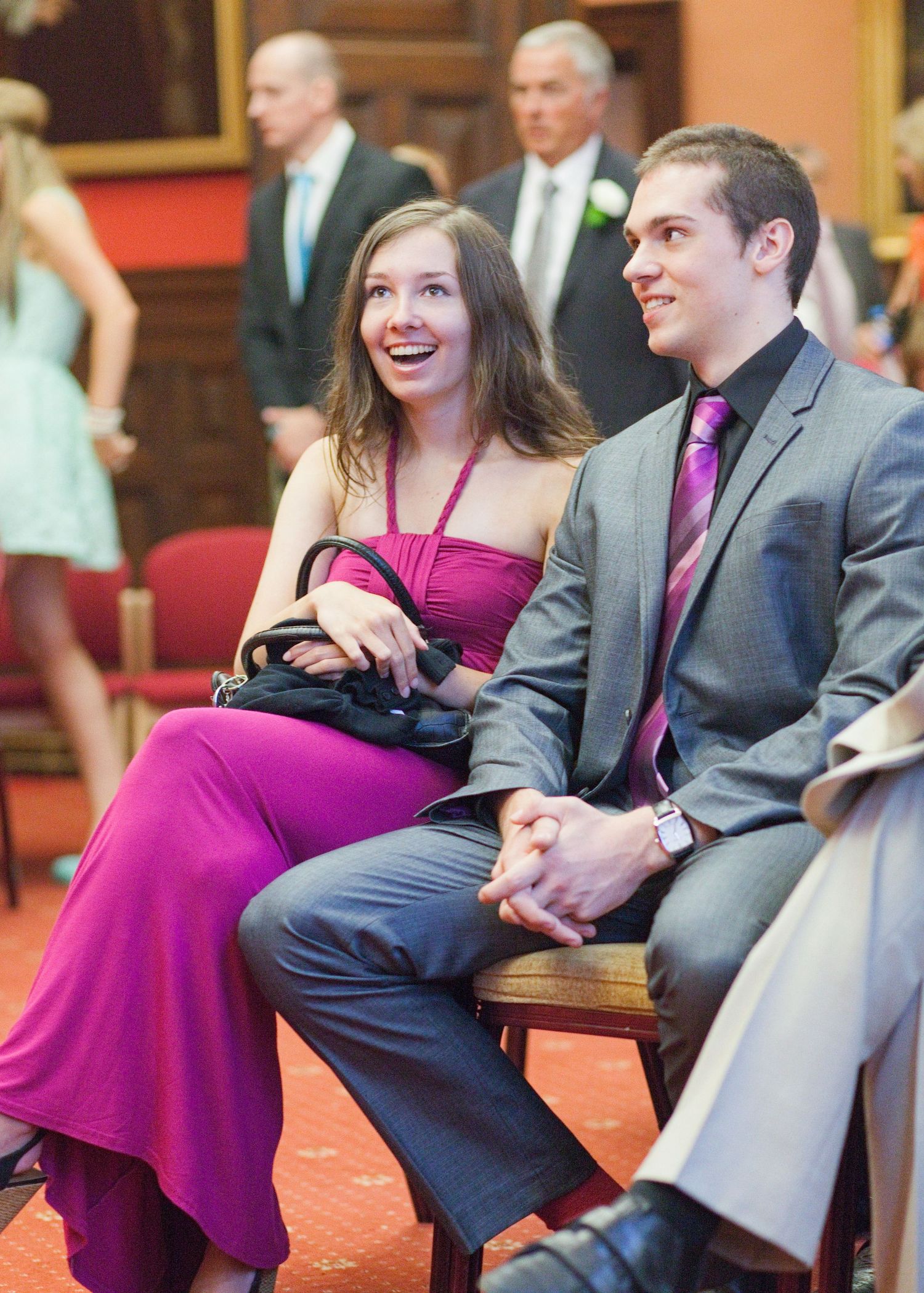 A couple in formal attire sits together on a bench, with the woman wearing a vibrant magenta dress.