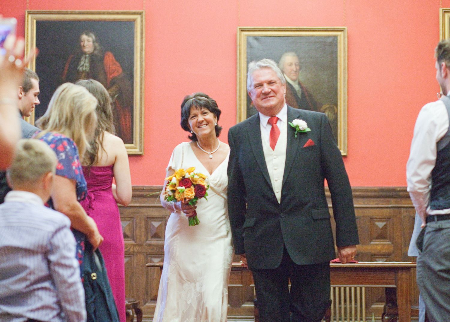 A couple smiling while walking together at their wedding ceremony in front of red walls with portraits.