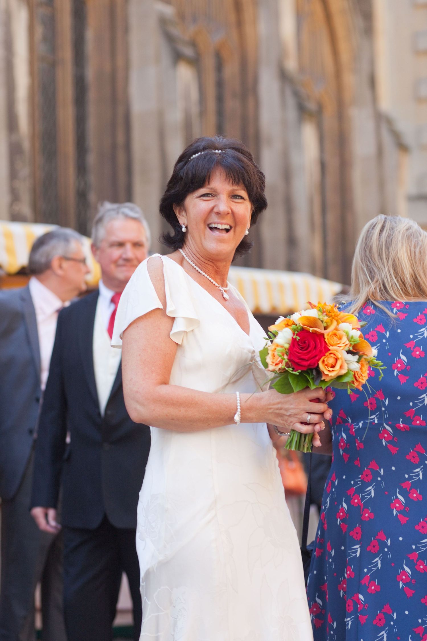 A bride in a white dress laughs joyfully while holding a vibrant orange and red bouquet outside a church.
