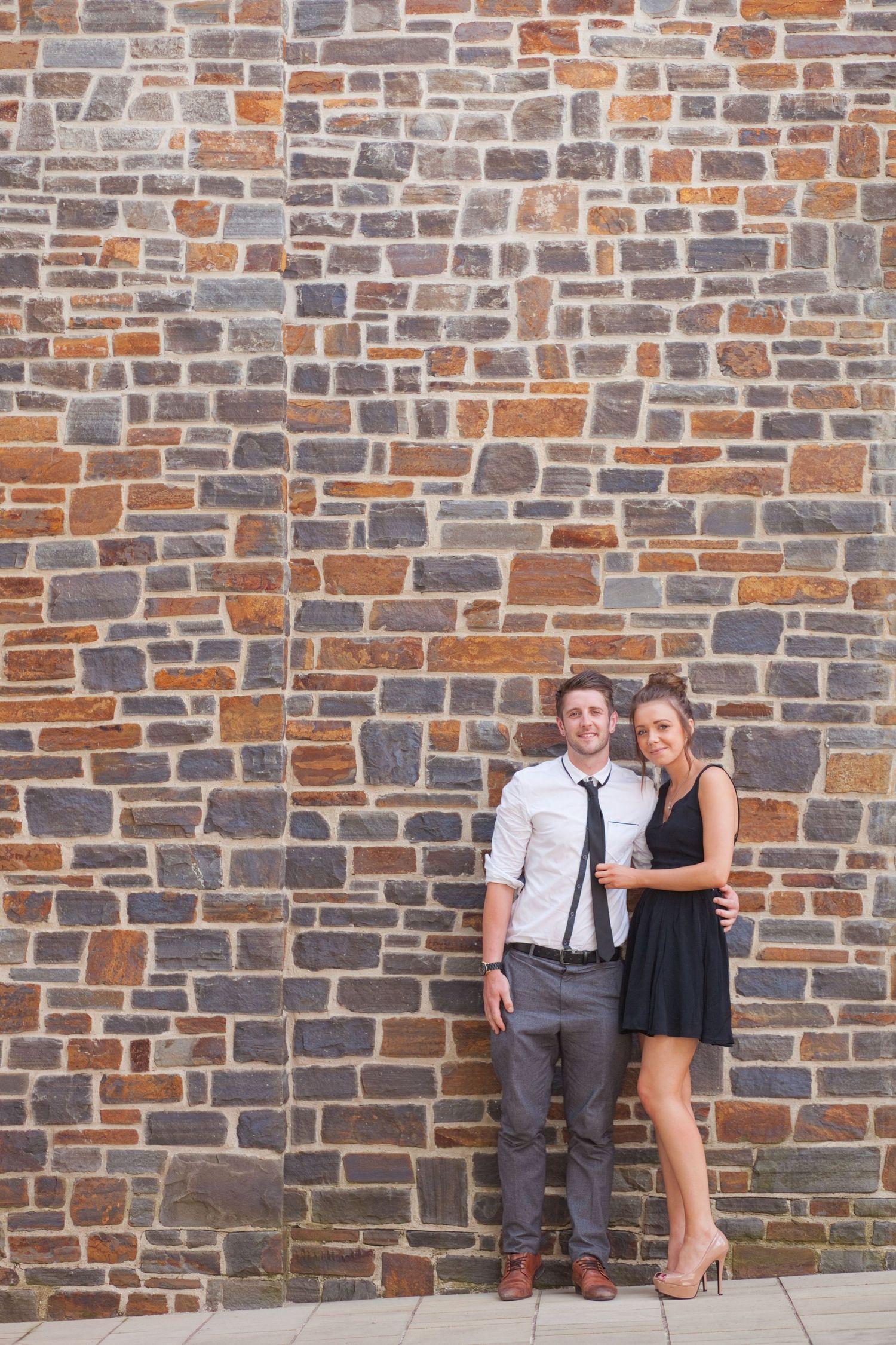 A couple poses casually against a rustic stone brick wall during a photo session.
