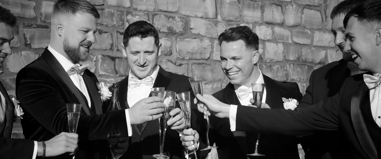 Groomsmen share drinks and laughs in a candid black and white moment.