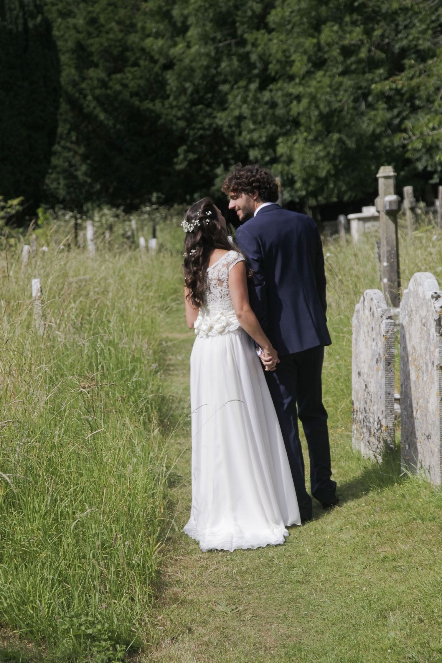A couple in wedding attire stands among tall grass and gravestones.