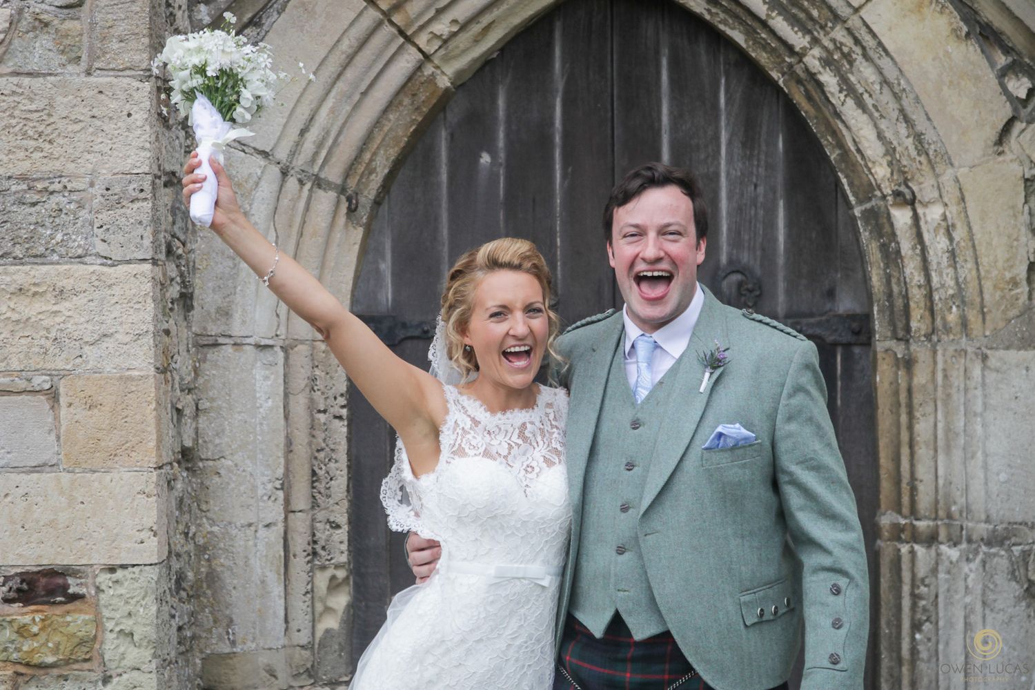 Wedding couple poses under stone archway of historic church building.