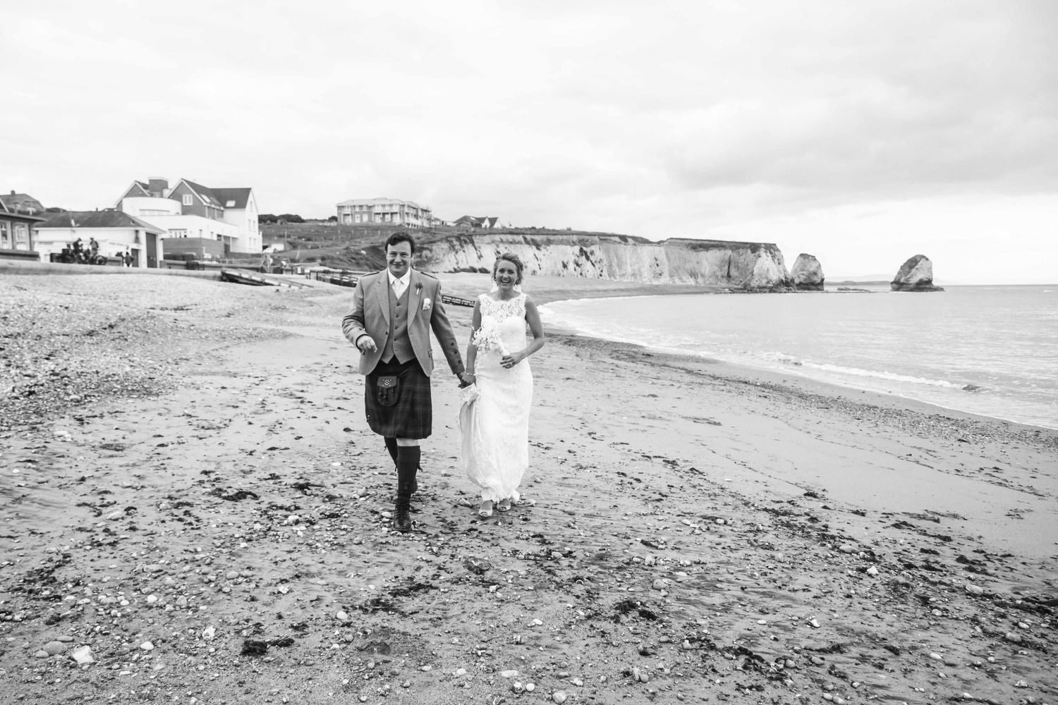 Black and white panoramic view of a couple walking along a pebbled beach near coastal cliffs.
