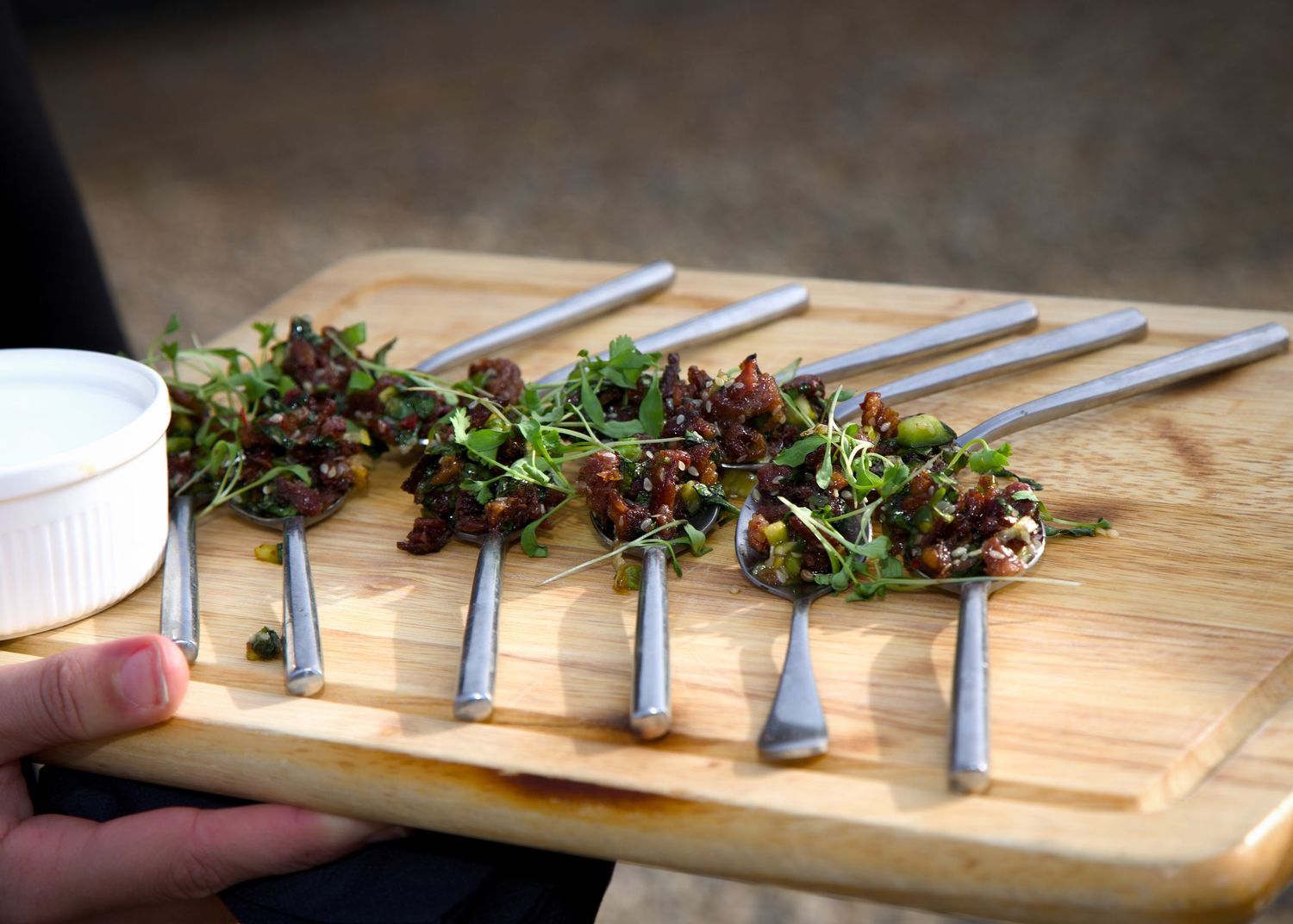 Small portions of microgreens arranged on silver spoons on a wooden serving board.