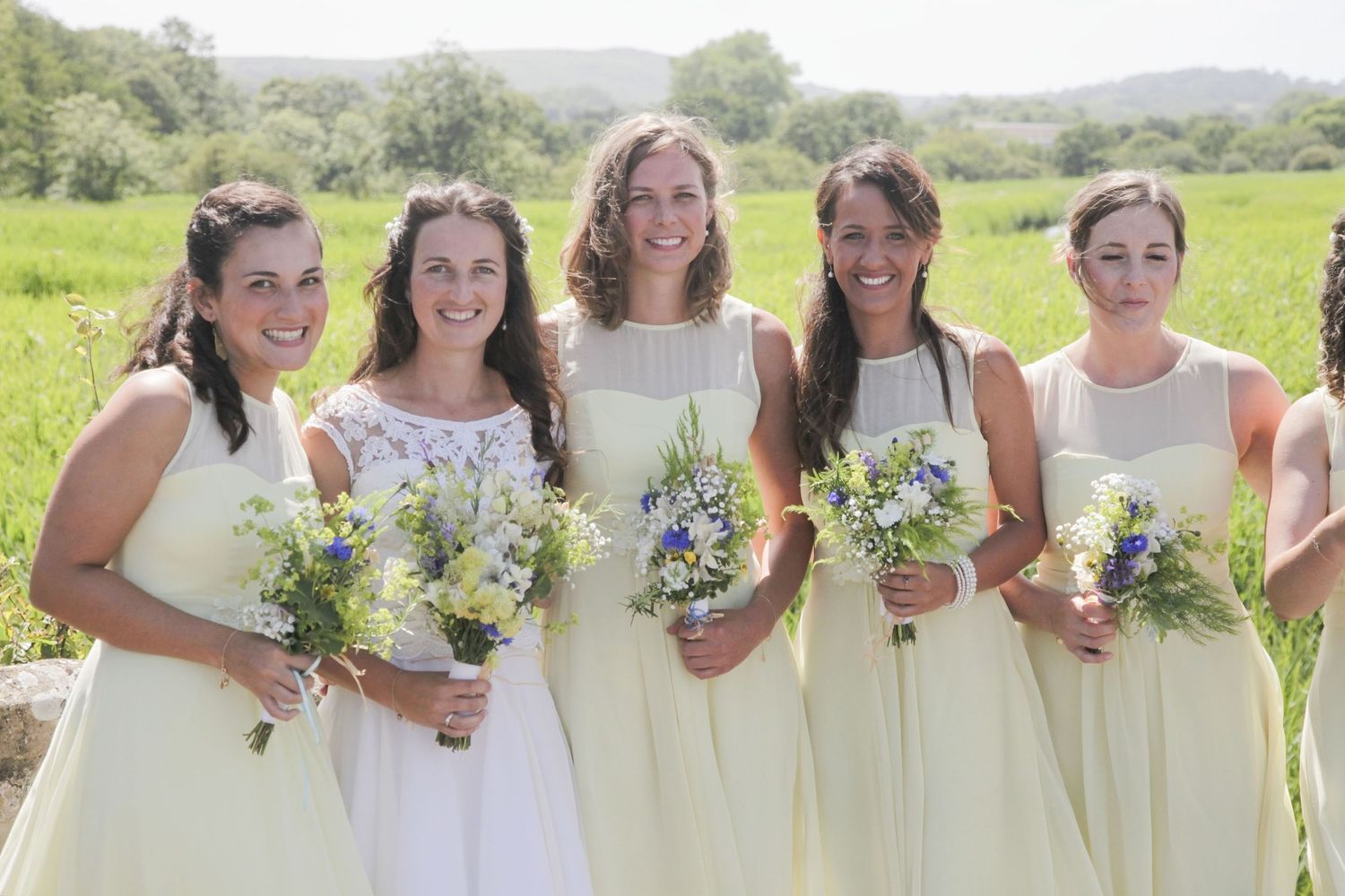 A line of bridesmaids in cream dresses hold wildflower bouquets against a pastoral landscape.
