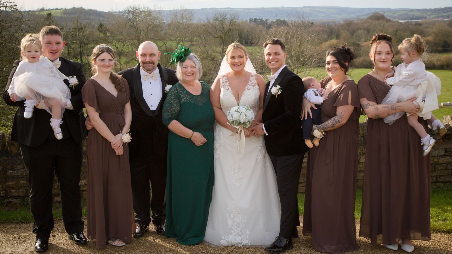 Wedding party in brown and green attire posing together against a scenic mountain backdrop.
