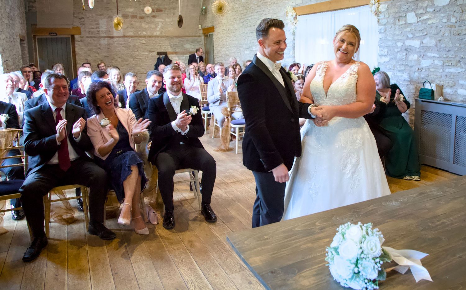 A wedding ceremony takes place in a rustic stone venue with guests watching the couple exchange vows at Folly Farm Somerset wedding.