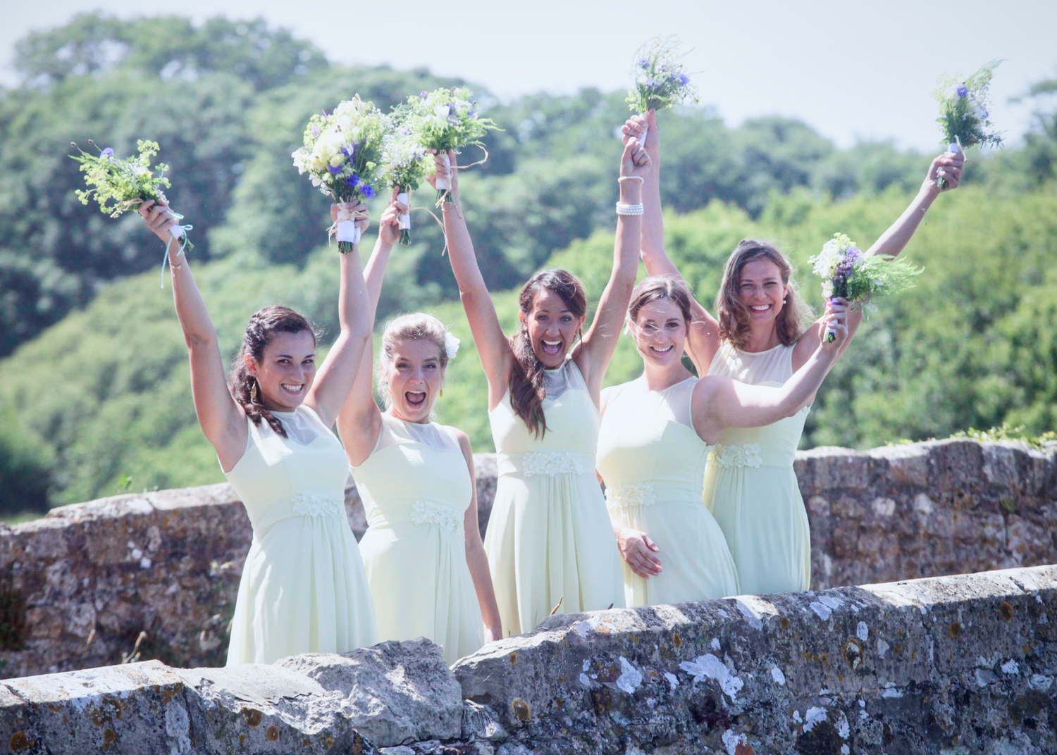 Bridesmaids in pale dresses celebrate on a stone wall holding bouquets overhead.