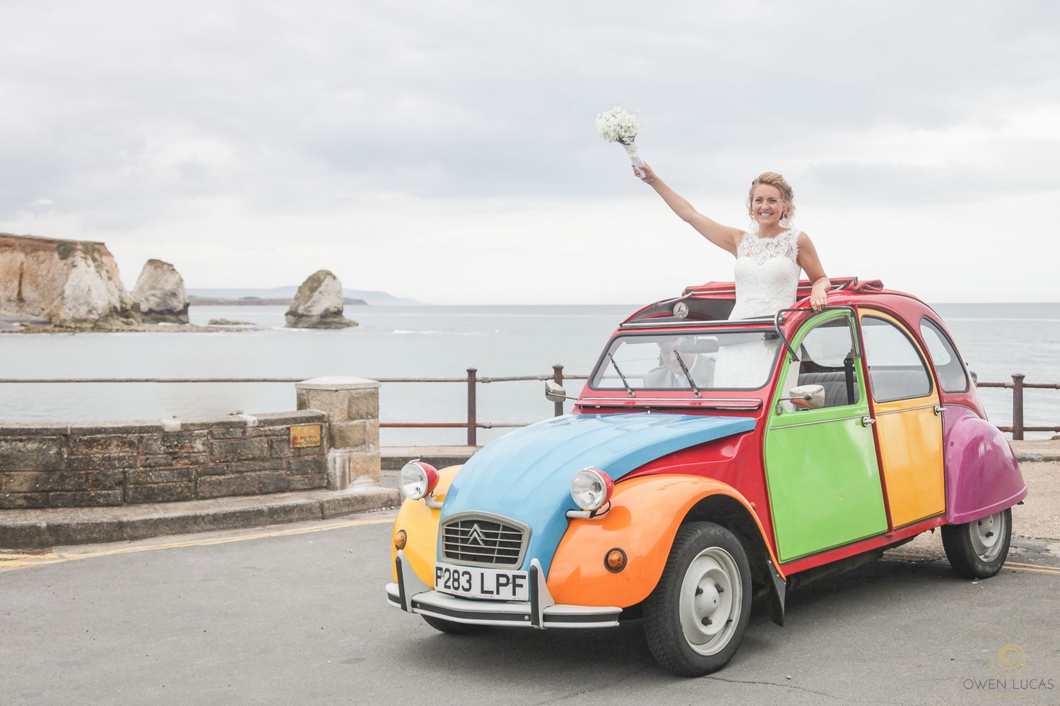 Person celebrates in colorful vintage Citroen 2CV car near coastal cliffs.