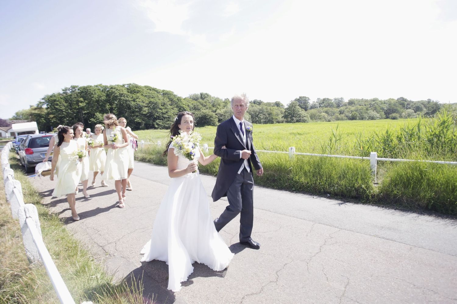 Wedding procession walking along a paved pathway with scenic marshland views.