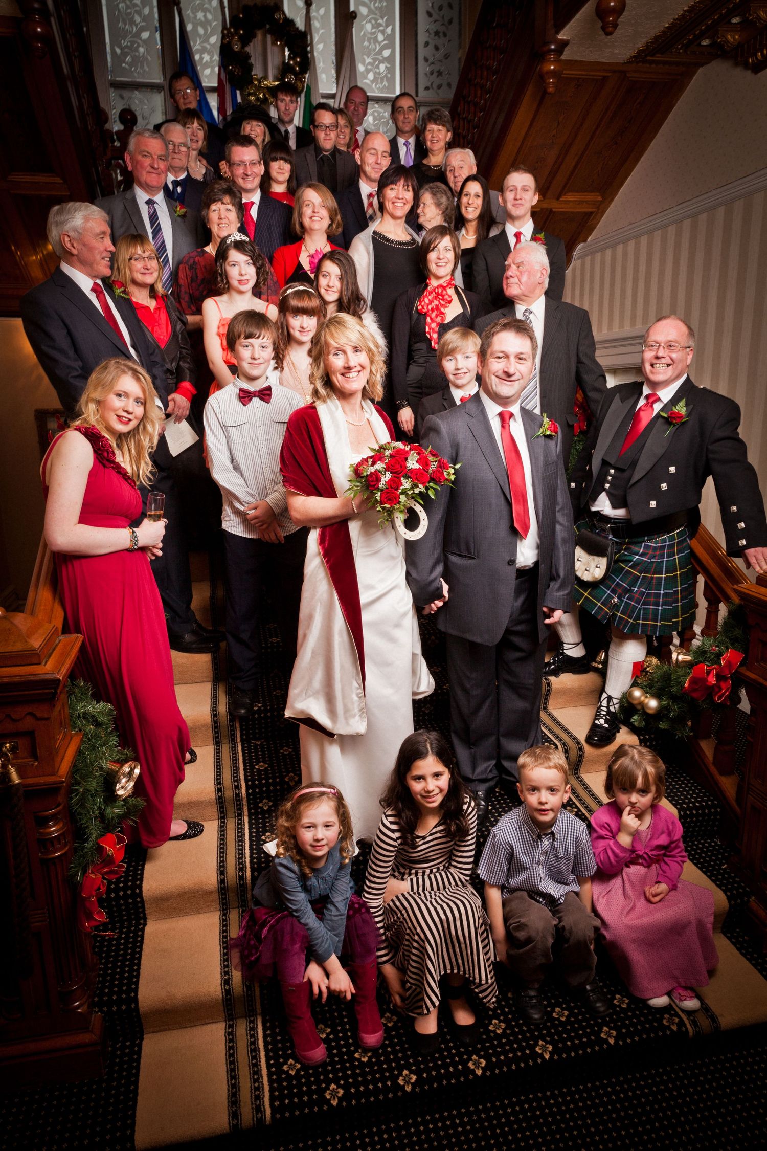 Large wedding party group photo on stairs with traditional Scottish kilts and red accents.
