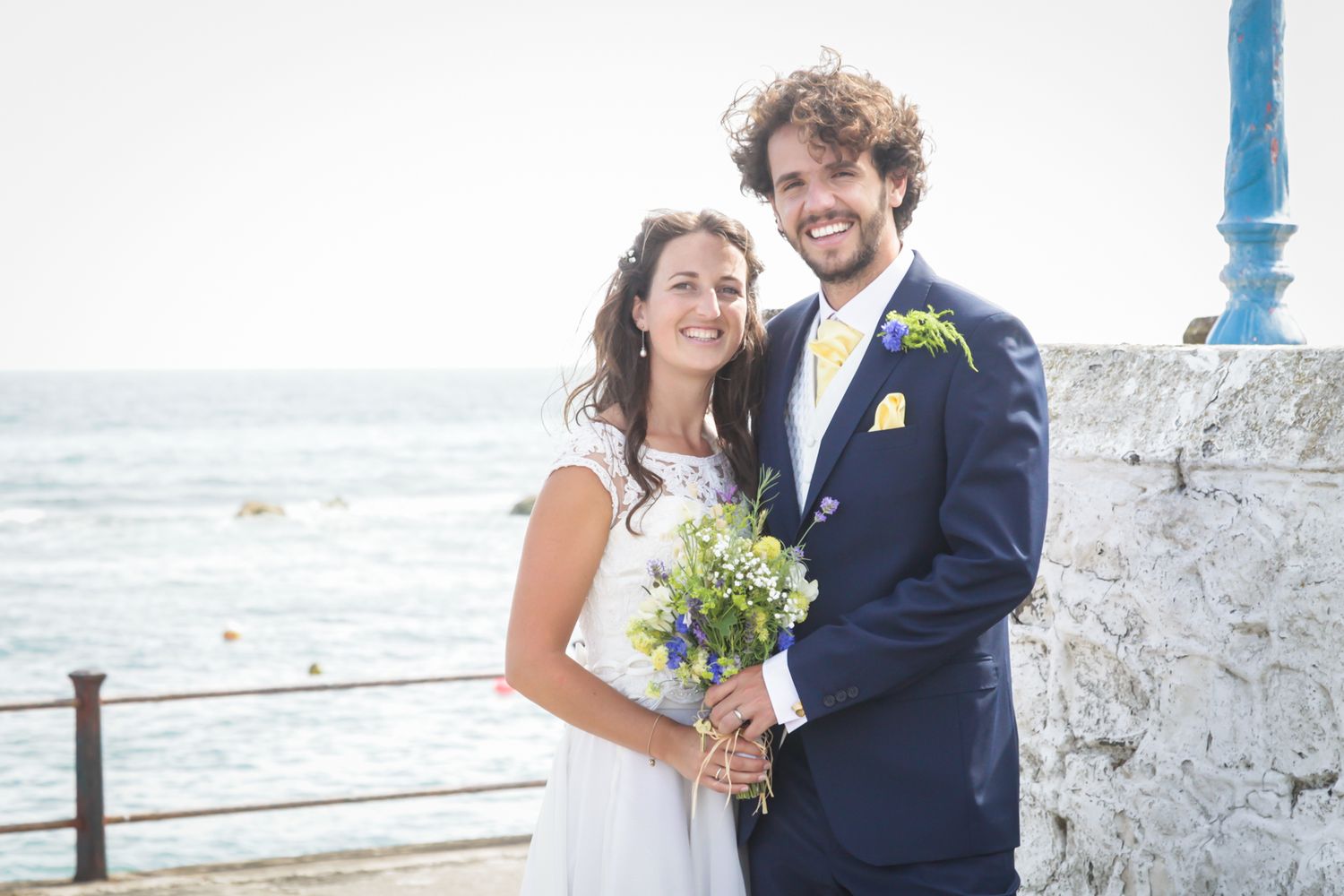 A newlywed couple poses by the ocean with the bride holding a yellow and white bouquet.