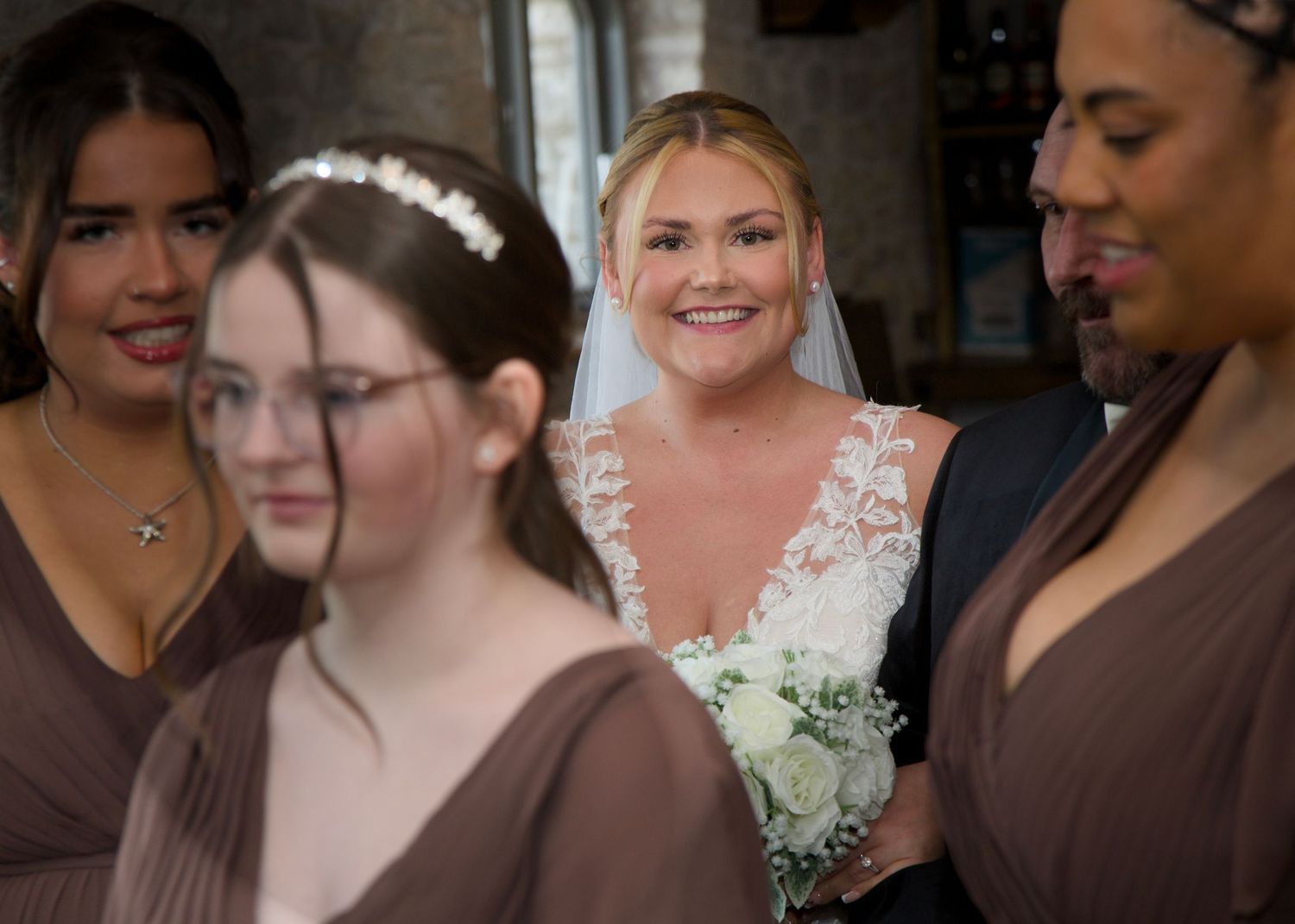 Bridesmaids in brown dresses stand together holding white bouquets at a wedding.