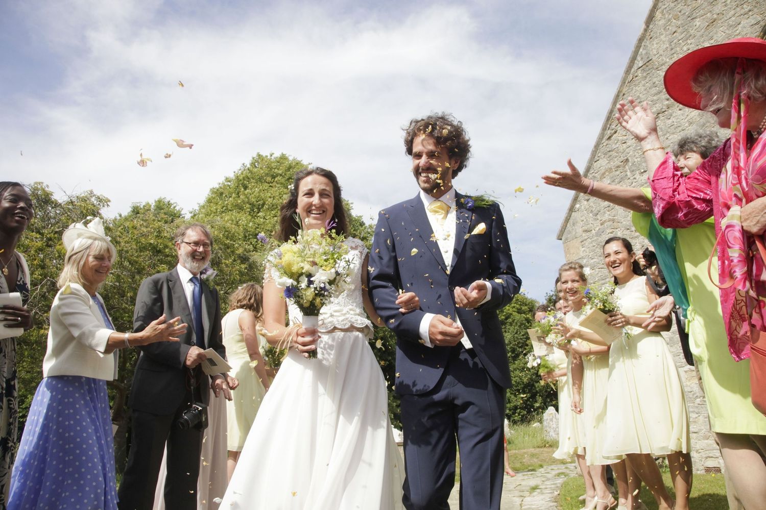 Wedding guests and party members gather for outdoor photos near a stone building.