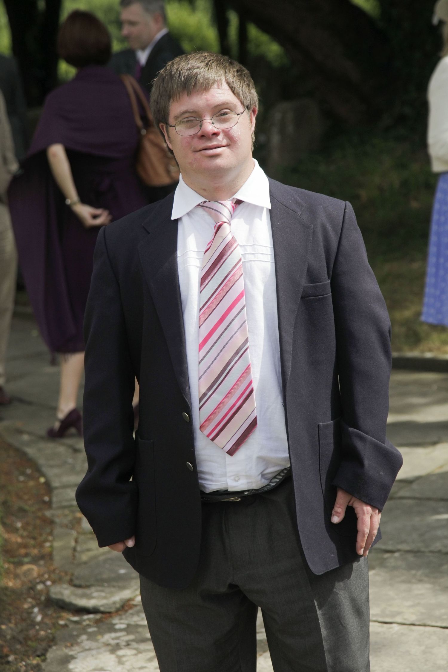 Formal wedding attendee in a dark suit and striped tie stands outdoors.