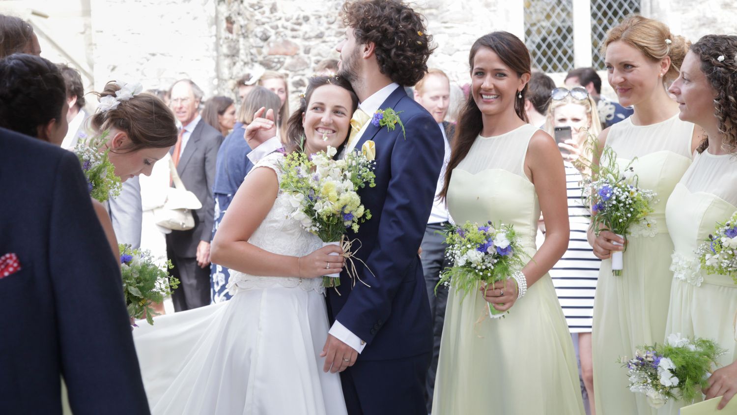 Wedding party members gather outdoors holding bouquets of white and green flowers.