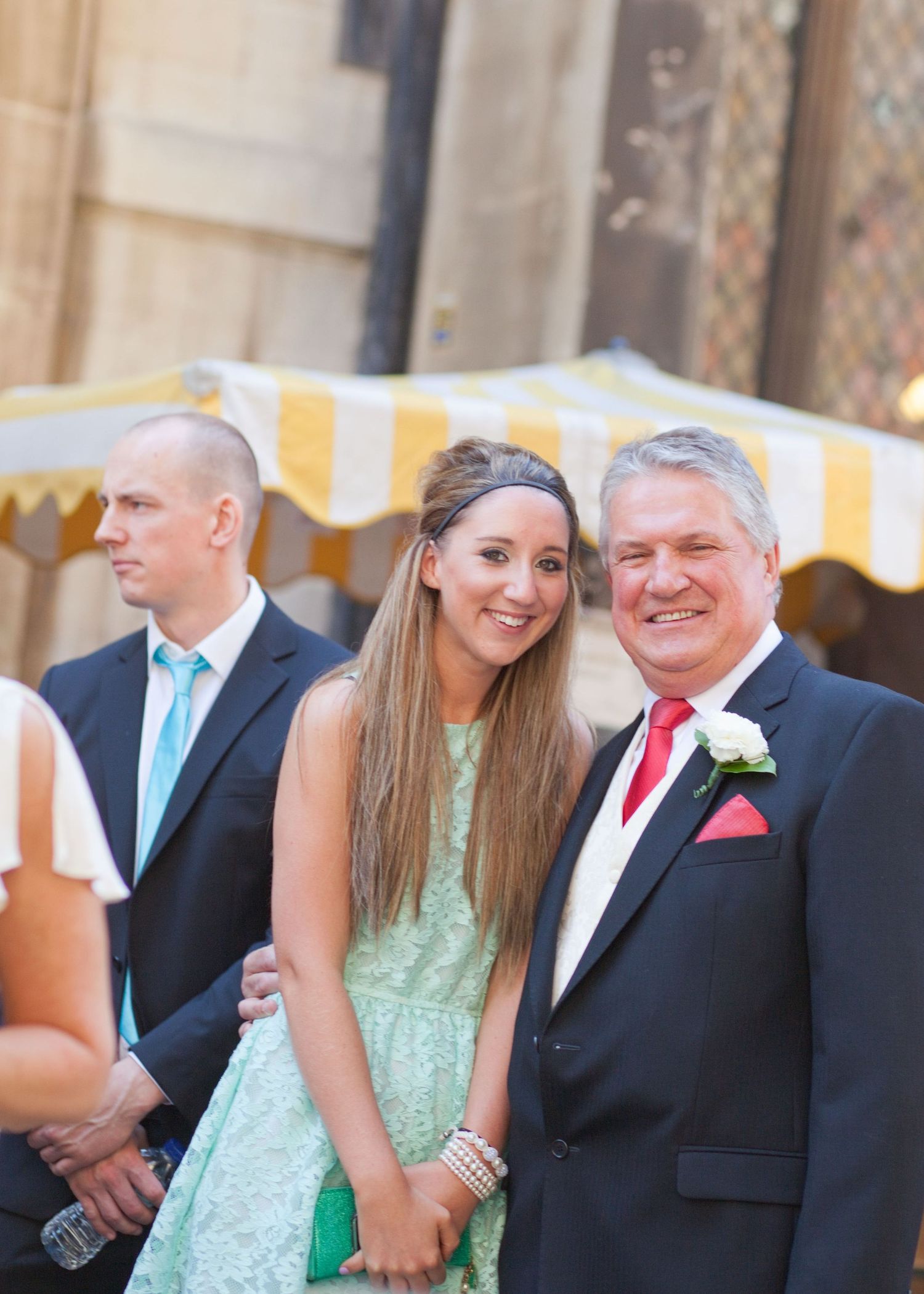 Wedding party members pose in formal attire against stone building backdrop.