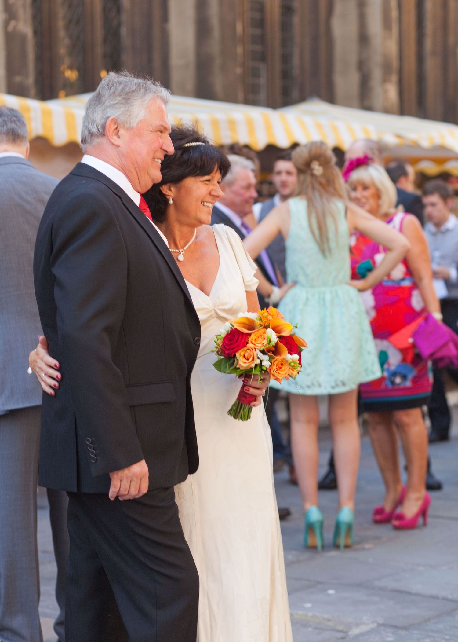 A wedding couple poses outside a historic church with guests in colorful dresses visible in the background.